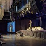 Robert Pearl on stage at Hint Summit in Denver, Colorado. Two large projection screens flank the speaker, one with a logo for the event; the other a live video feed